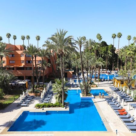 Kenzi Rose Garden Hotel Marrakesh Buitenkant foto The photo shows a resort-style area with several swimming pools surrounded by palm trees. The pools are clear blue and are lined with numerous lounge chairs for relaxation. There is a vibrant atmosphere, highlighted by lush greenery and a sunny sky. 