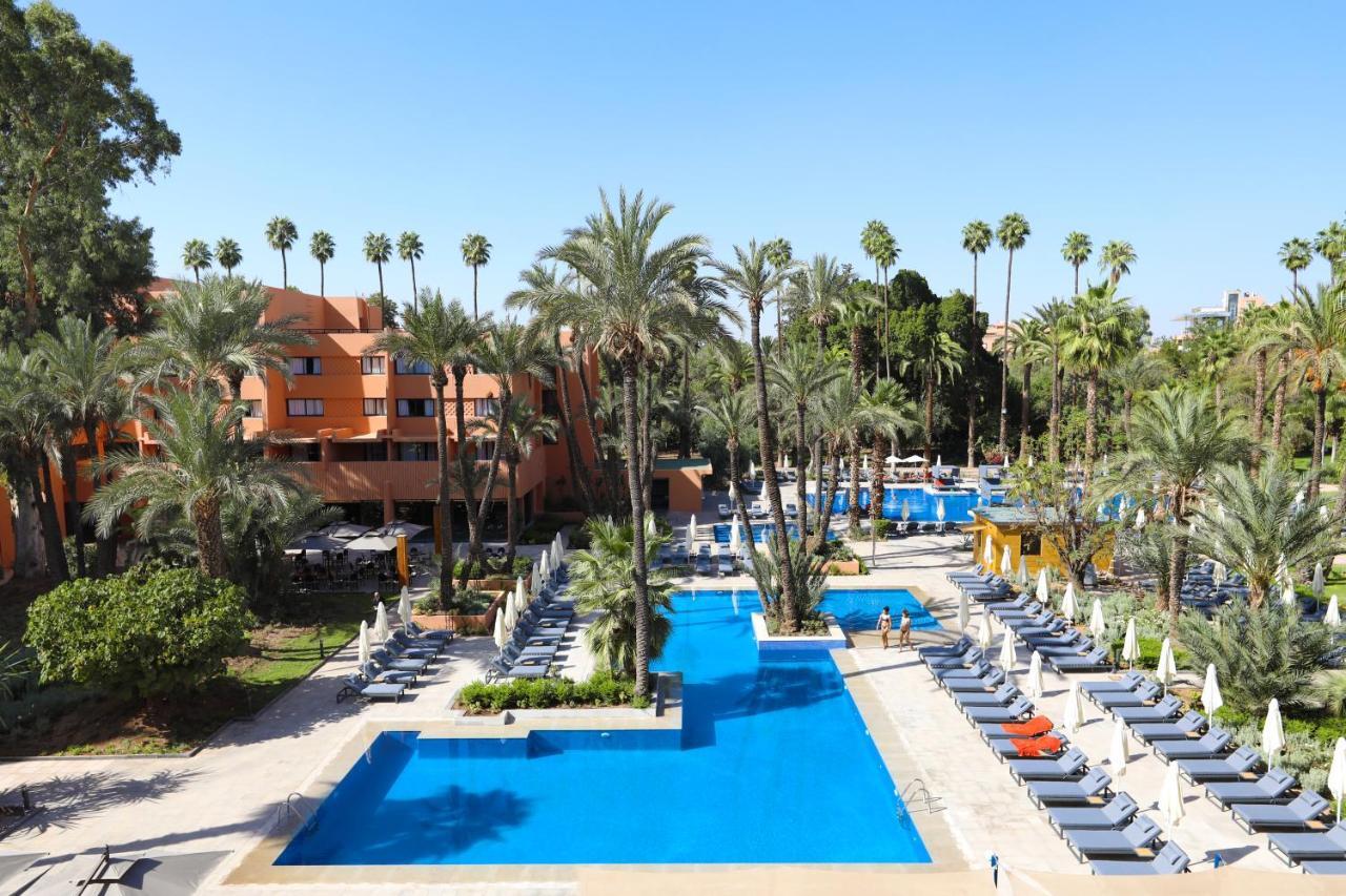 Kenzi Rose Garden Hotel Marrakesh Buitenkant foto The photo shows a resort-style area with several swimming pools surrounded by palm trees. The pools are clear blue and are lined with numerous lounge chairs for relaxation. There is a vibrant atmosphere, highlighted by lush greenery and a sunny sky. 