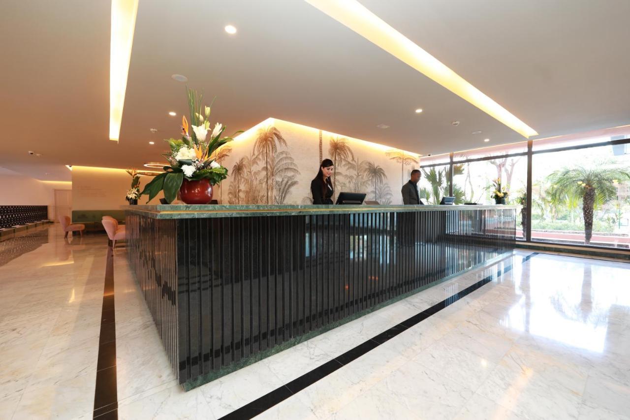 Kenzi Rose Garden Hotel Marrakesh Buitenkant foto The image shows a modern reception area, likely for a hotel or office building. There is a stylish front desk made of dark materials with a smooth surface. Behind the desk, two staff members are engaged in their work, possibly checking in guests or a